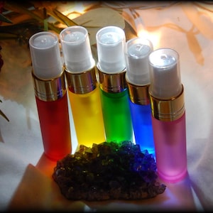 May include: Five colorful spray bottles in red, yellow, green, blue, and pink, each with a gold band and white top. A cluster of dark, multi-colored crystals sits in front of the bottles. The bottles are arranged in a semi-circle.