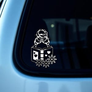 Może przedstawiać: Biała naklejka winylowa przedstawiająca kobietę z włosami w koku, czytającą książkę. Książka ma serca i kwiatowy wzór. Naklejka jest otoczona kwiatami.