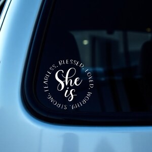 以下が含まれることがあります： 白いビニール製の車の窓用ステッカー。筆記体で「She is」と書かれ、「Fearless, Blessed, Loved, Worthy, Strong」という言葉が円形に配置されています。