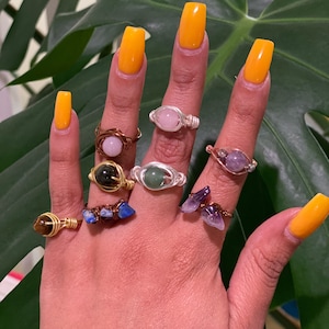 Könnte beinhalten: Eine Hand zeigt eine Sammlung von drahtgewickelten Edelsteinringen. Die Ringe weisen verschiedene Steine in Rosa-, Grün-, Lila- und Blautönen auf, die in Silber- und Kupferdraht gefasst sind. Die Nägel sind leuchtend gelb lackiert.