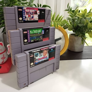 May include: Three Super Nintendo Entertainment System (SNES) game cartridges stacked on top of each other. The top cartridge is NHLPA Hockey 93, the middle cartridge is PGA Tour Golf, and the bottom cartridge is NFL Football.