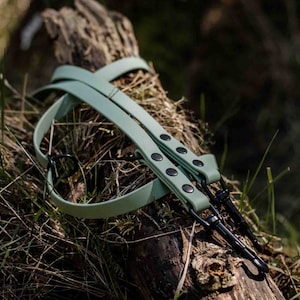 May include: A light green camera strap with black metal hardware and black button accents. The strap is lying on a log in a forest setting.