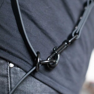 May include: A black leather strap with a black metal clasp. The strap is worn over a person's shoulder and across their chest.