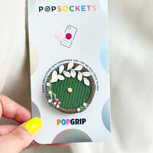 May include: A green and brown phone grip with a white door and leaves. The grip is designed to look like a hobbit hole. The packaging says "PopGrip" and "POPSOCKETS".