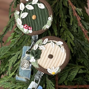May include: Two round, brown, wooden-looking, decorative,  retror-style door-shaped,  keychain badge holders with green and white floral accents. The top holder has a green door with a gold knob and a red and white mushroom. The bottom holder has a white door with a brown knob and a white daisy.