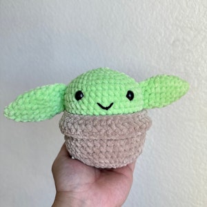 Op de afbeelding: Een handgemaakt gehaakt pluche speelgoed van een personage met grote oren, een groene kop en een bruin lichaam. Het speelgoed heeft zwarte ogen en een kleine glimlach. Het pluche wordt in een hand tegen een witte achtergrond gehouden.