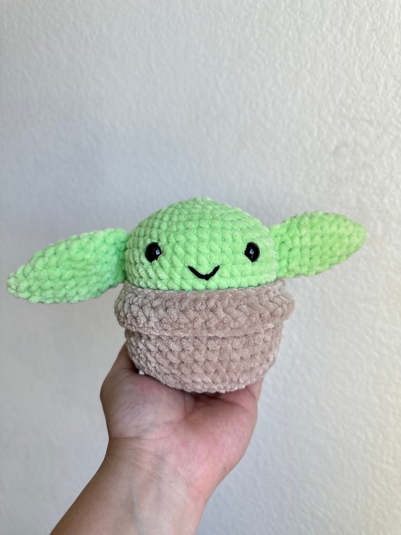 Op de afbeelding: Een handgemaakt gehaakt pluche speelgoed van een personage met grote oren, een groene kop en een bruin lichaam. Het speelgoed heeft zwarte ogen en een kleine glimlach. Het pluche wordt in een hand tegen een witte achtergrond gehouden.