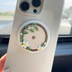 May include: A white phone case with a round, white, brown, and green decorative sticker featuring a small, wooden door with a vine and flowers.
