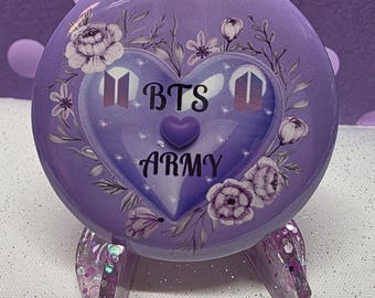 May include: A round, purple decorative item with the text "BTS ARMY" inside a heart shape, surrounded by floral accents. The item is supported by a glittery stand. The design features the BTS logo and a small heart.
