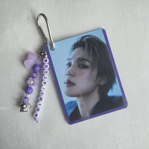 May include: A rectangular photo card featuring a person with dark hair and a purple border. The card is attached to a silver carabiner clip with a beaded charm. The charm includes a purple bow, beads, and the name "BTS JIMIN" spelled out.