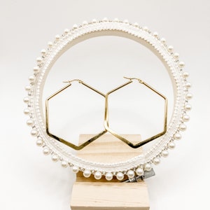 May include: A pair of gold hexagonal hoop earrings displayed on a white pearl-embellished jewelry stand.