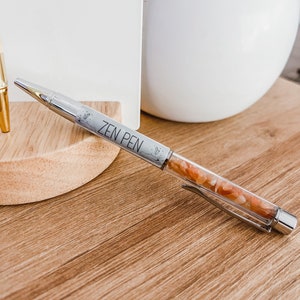 May include: A silver pen with a clear barrel filled with small orange and white stones. The pen has the words "ZEN PEN" printed on the side.