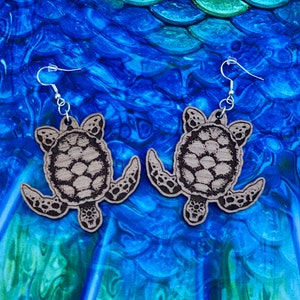 Peut inclure: Une paire de boucles d'oreilles en bois en forme de tortues de mer. Les boucles d'oreilles sont brunes avec un grain de bois clair et ont des crochets argentés.