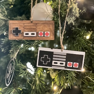 May include: Two Christmas tree ornaments shaped like classic video game controllers. One is made of wood with black and red details, the other is black and white. Both are hanging from twine strings.