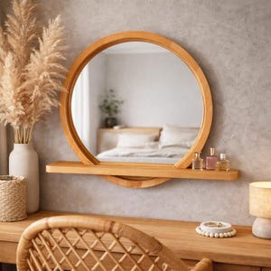 Op de afbeelding: Een ronde houten spiegel met een plank, die een slaapkamer weerspiegelt. Drie parfumflesjes staan op de plank. Een vaas met droogbloemen en een gevlochten mand staan links. De spiegel heeft een diameter van ongeveer 60 cm.
