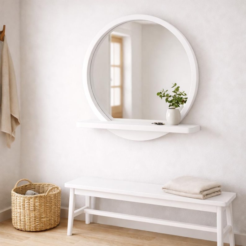 Op de afbeelding: Een witte, ronde spiegel met een plank, een kleine witte vaas met groen en een witte bank. Een geweven mand en een opgevouwen handdoek zijn ook zichtbaar. De spiegel is aan een witte muur bevestigd.
