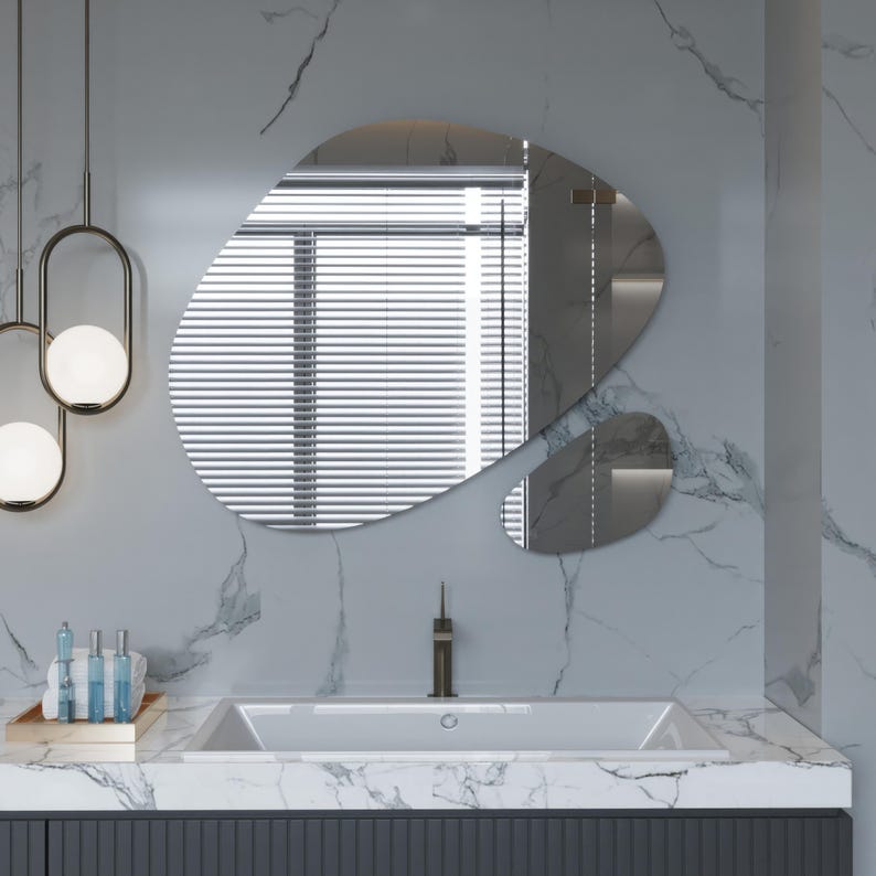 Pu&ograve; includere: Un lavabo in porcellana bianca con un rubinetto unico in un bagno moderno con un piano in marmo bianco e uno specchio di forma organica.