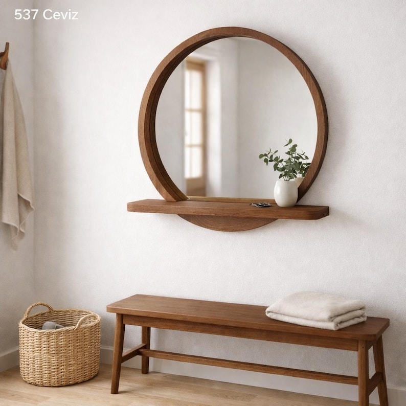Op de afbeelding: Een ronde houten spiegel met een plank, een kleine witte vaas met groen en een houten bank. De spiegel is aan een witte muur bevestigd. Een gevlochten mand en een opgevouwen deken zijn ook zichtbaar.