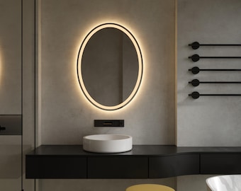 Puede incluir: Un tocador de baño moderno con una encimera negra y un lavabo ovalado blanco. Un espejo ovalado grande con un anillo de luz LED blanco está montado en la pared sobre el lavabo. Un taburete amarillo está en primer plano.