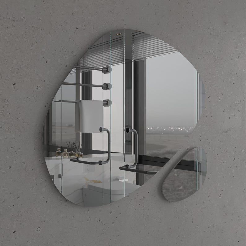 Pu&ograve; includere: Uno specchio da bagno moderno con una forma irregolare e organica. Lo specchio riflette una vista di un bagno con un portasciugamani, una finestra e un lavandino.