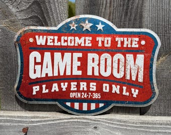 May include: A rectangular metal sign with a red, white, and blue color scheme, mounted on a wooden fence. The sign reads "WELCOME TO THE GAME ROOM PLAYERS ONLY" in white text. The sign has a distressed, vintage appearance.