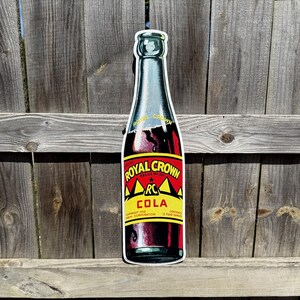 May include: Vintage Royal Crown Cola metal sign featuring a red and yellow bottle with the text "Royal Crown Cola" and "RC" in a star. The sign is a reproduction of an old-fashioned soda bottle.