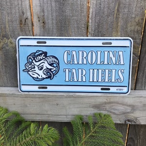May include: A blue and white metal license plate with the text "CAROLINA TAR HEELS" and a cartoon ram mascot with the text "UNC" on its hat.