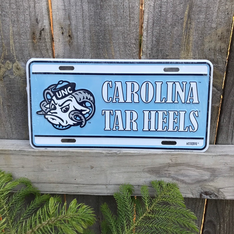 May include: A blue and white metal license plate with the text "CAROLINA TAR HEELS" and a cartoon ram mascot with the text "UNC" on its hat.