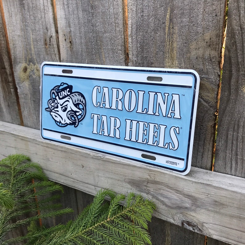 May include: A blue and white metal sign with the text "Carolina Tar Heels" and a cartoon mascot of a ram with a blue and white hat. The sign has a UNC logo on the mascot.