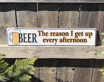 May include: White metal sign with black trim and orange text. The sign reads "The reason I get up every afternoon BEER" and features a cartoon image of a beer mug.