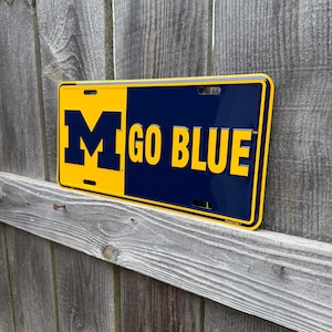 May include: A rectangular license plate in yellow and navy blue. The left side has a large navy blue "M", and the right side displays "GO BLUE" in navy blue text. The plate is mounted on a weathered wooden fence.