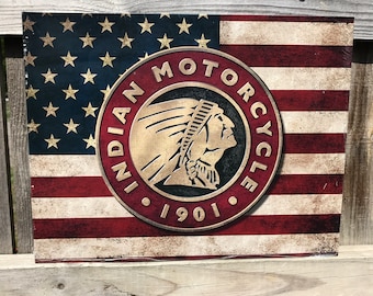Op de afbeelding: Een rechthoekig bord met een verweerde Amerikaanse vlag. Het bord heeft een cirkelvormig embleem met de tekst "Indian Motorcycle 1901" en een profiel van een indiaanse chef. Het bord is gemonteerd op een houten hek.