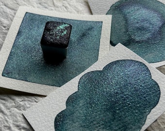 May include: Three watercolor paint samples on white paper. The samples are a dark teal green with a shimmery finish. The paint is in a square pan with a black lid.