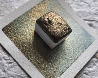 May include: A small, square, metallic gold watercolor pan with a white plastic casing. The pan is sitting on a white card with a metallic gold and green wash.