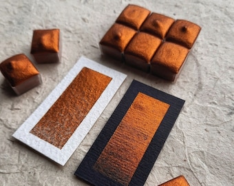 May include: A set of six metallic copper watercolor paint pans, arranged in a rectangle, with two additional pans on a white and black background. The pans are arranged in a grid pattern, with each pan having a slightly different shade of copper.
