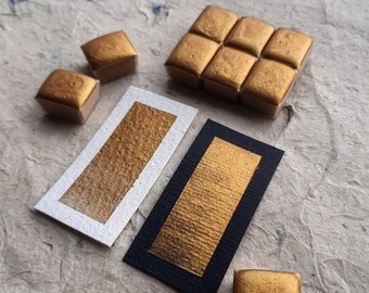 May include: A collection of metallic gold watercolor pans and two rectangular paper swatches. One swatch features a gold paint stroke on white paper, while the other displays a gold paint stroke on black paper, showcasing the pigment's opacity.