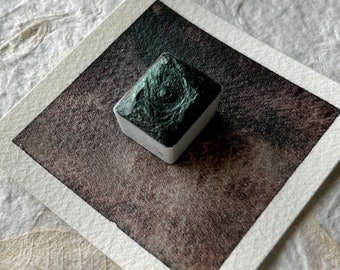 May include: A dark green watercolor paint pan with a metallic sheen. The paint is in a square pan with a white label. The pan is sitting on a white surface.