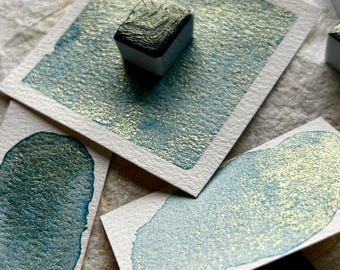 May include: Close-up of watercolor paints and swatches. The paints are in small, white rectangular containers with dark green tops. The swatches are on small white cards, showing a teal and gold metallic sheen. The background is a textured, off-white surface.