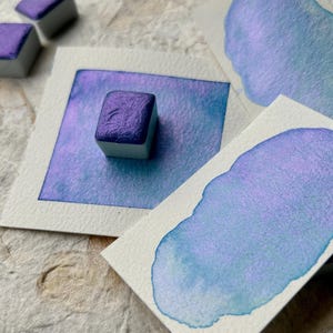 以下が含まれることがあります： 白い紙の上に、紫色の光沢のあるものが1つ入った、水彩絵の具の皿が3つ置かれています。皿は、紫色と青色の水彩のぼかしの上に置かれています。