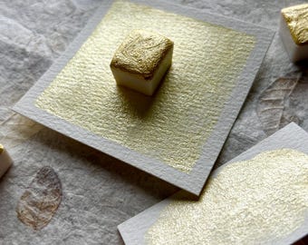 May include: Close-up of gold watercolor paints and swatches. The paints are small, square, and have a gold metallic finish. The swatches are on white paper, showcasing the paint's shimmering quality. The background is a textured, neutral-toned surface.