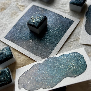 Puede incluir: Primer plano de pinturas de acuarela y muestras de color. Las pinturas son de color verde azulado oscuro con motas doradas, y las muestras muestran un degradado de verde azulado y plata. Las pinturas est&aacute;n en peque&ntilde;os recipientes cuadrados.