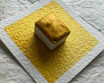 May include: A small, square, white pan with a gold metallic paint inside. The pan is sitting on a white textured surface.