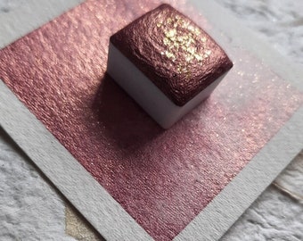 May include: A square watercolor paint pan with a metallic rose gold hue, accented with gold flecks, sits on a matching rectangular paper. The paint pan is white on the sides. The paper is set on a textured, off-white surface.