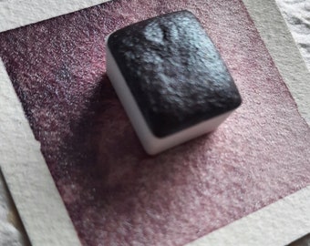 May include: A single watercolor pan in a white plastic tray. The pan is filled with a dark purple watercolor paint. The paint has a shimmery, metallic finish.