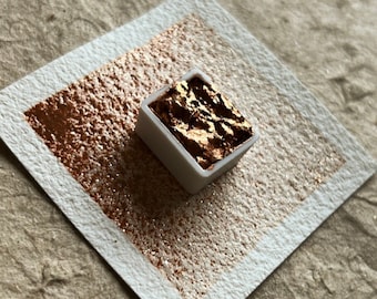 May include: A small square pan of metallic gold watercolor paint. The paint is a shimmering, chunky texture. The pan is sitting on a white piece of paper.