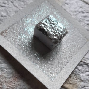 May include: A small, square, white watercolor pan with a silver shimmer. The pan is sitting on a white paper surface.