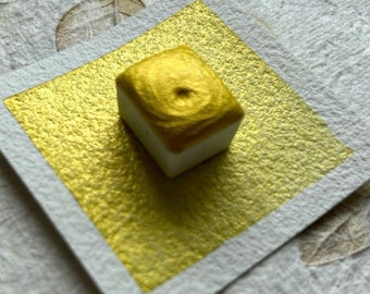 May include: A small, square, gold watercolor pan with a metallic sheen. The pan is sitting on a white textured paper.