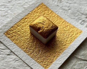 May include: A small, square, white pan of metallic gold watercolor paint. The pan is sitting on a white textured paper surface.
