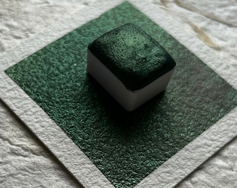 May include: A square pan of dark green watercolor paint with a metallic sheen. The pan is sitting on a white square of paper.
