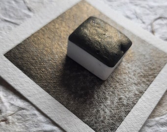 May include: A single square watercolor pan with a metallic black and gold finish. The pan is sitting on a white paper surface.